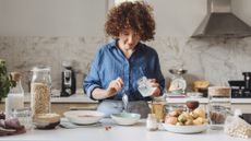 A woman with curly hair assembles overnight oats in a home kitchen. Her shirt is blue. Various igredients are laid out on the counter in front of her.