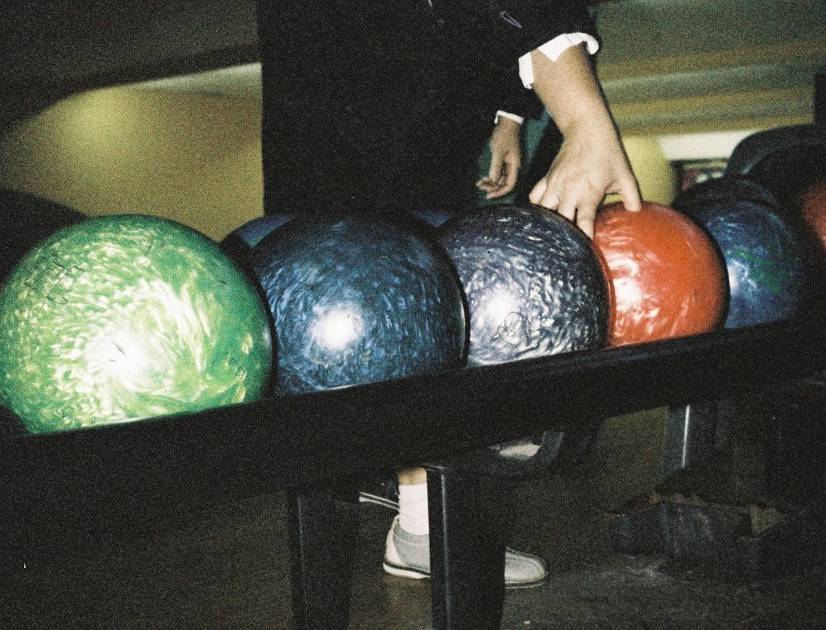 Hand selecting a red bowling ball from a rack filled with green, blue, and purple bowling balls, person wearing white sneakers