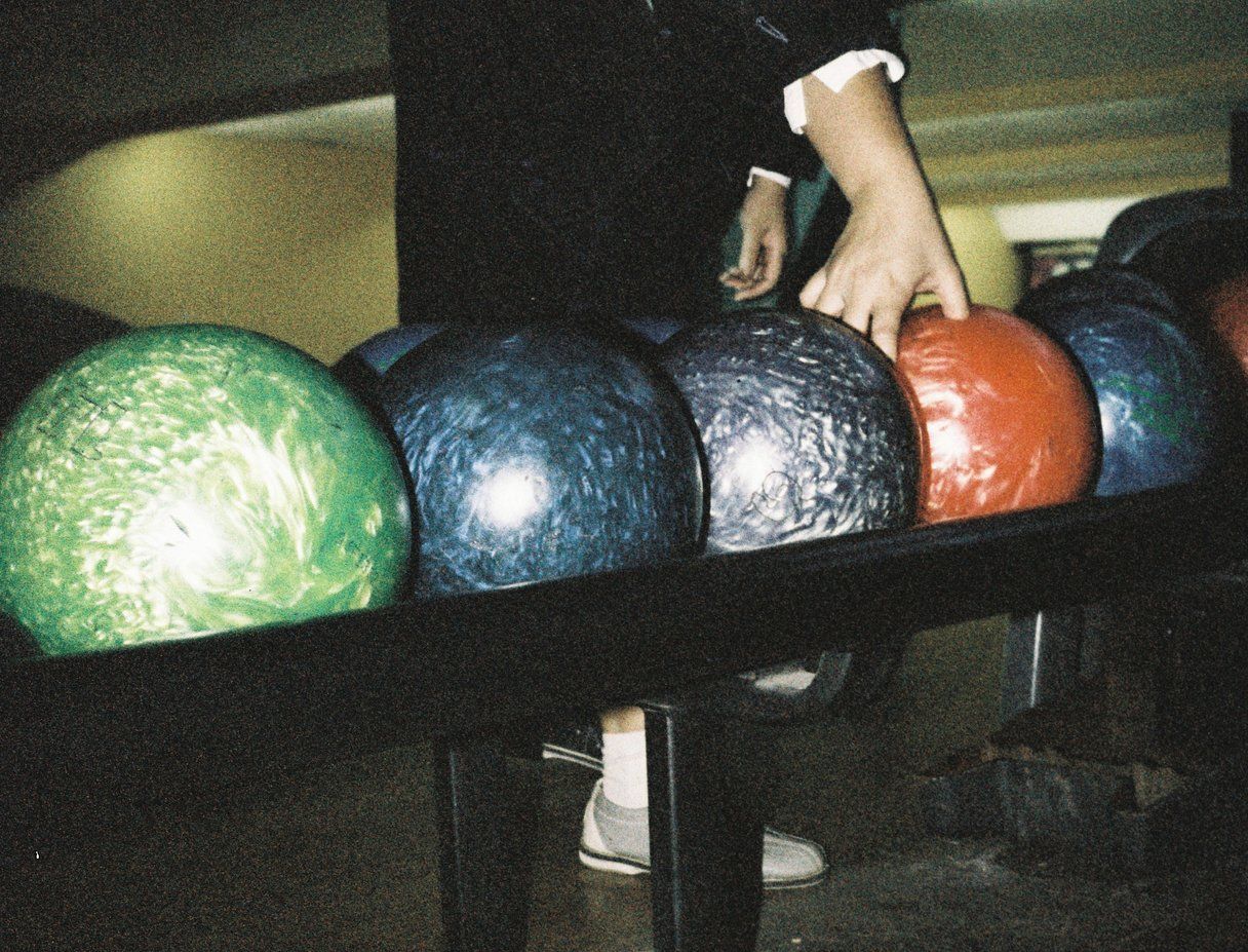 Hand selecting a red bowling ball from a rack filled with green, blue, and purple bowling balls, person wearing white sneakers