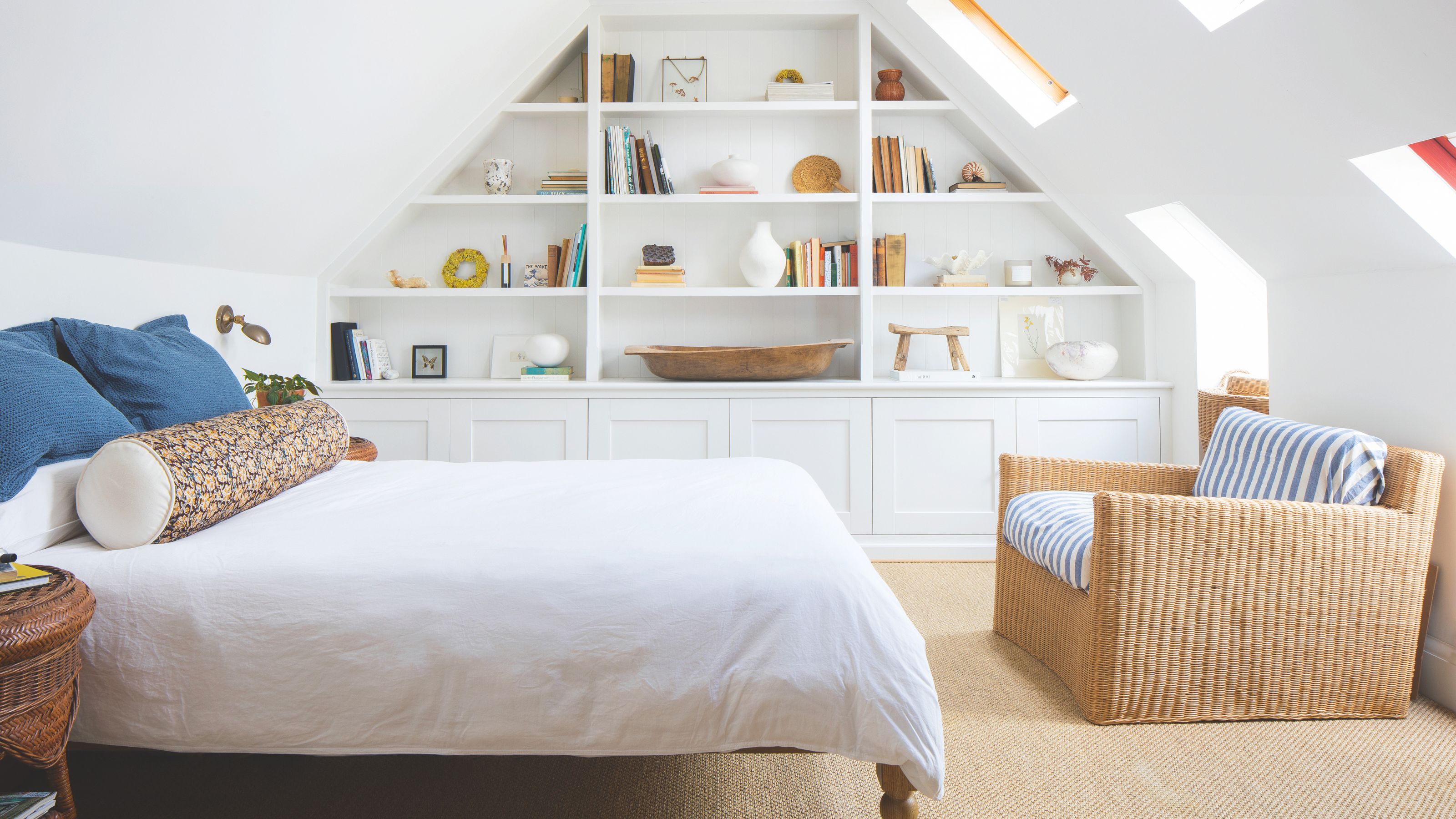 White painted loft bedroom with a white bed and a wicker chair at the end of it, and fitted shelves on the wall to the side of it