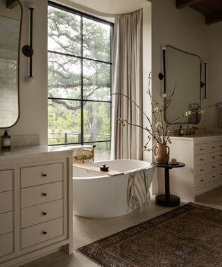 White bathroom with two sink vanity areas, large mirrors, freestanding bathtub, side table and patterned Persian style rug