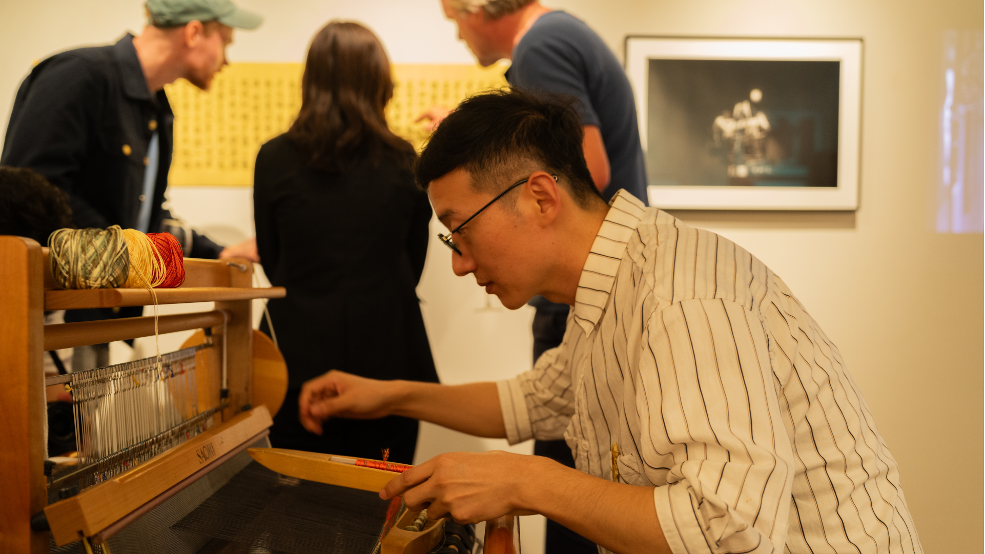 A man in a striped shirt demonstrates weaving on a small wooden loom, passing a shuttle of red thread through the warp, as visitors look at the artworks behind him.