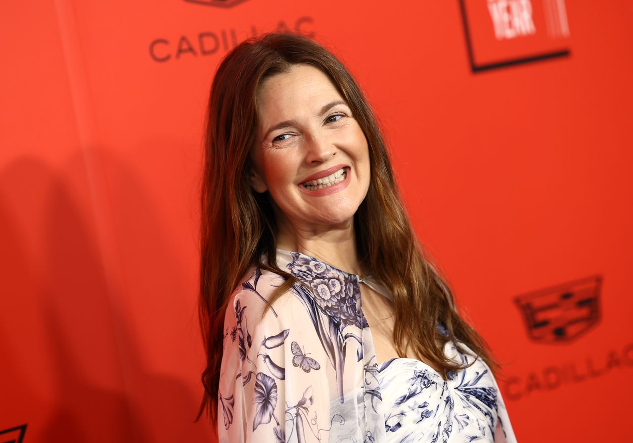 A headshot of Drew Barrymore on a red carpet smiling over her shoulder