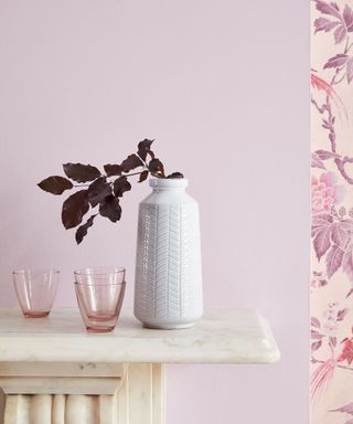 Close up of mauve painted wall above fireplace, floral purple wallpaper to the right, white vase with dried purple flowers, small pink tumbler glasses