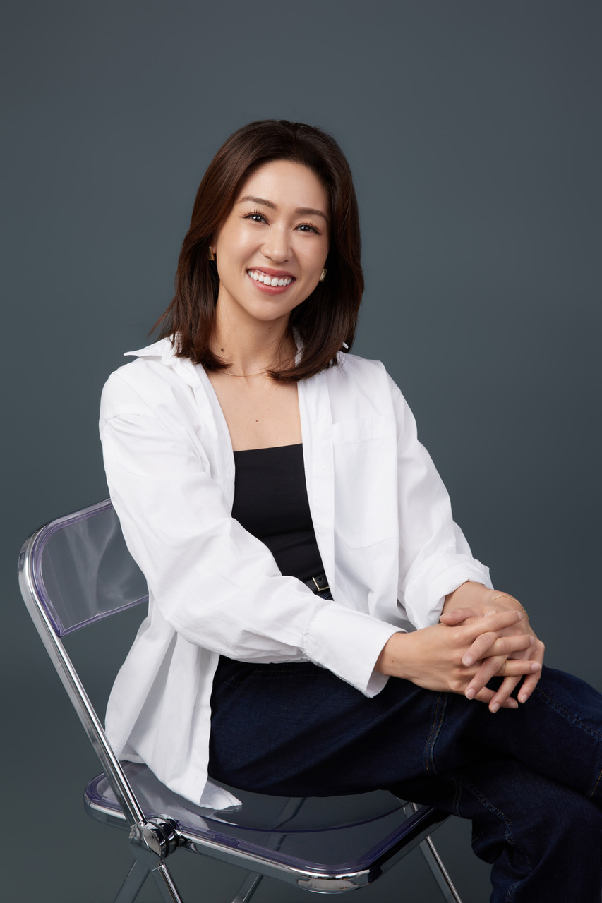 a young Asian woman in a black shirt, white overshirt and black pants sitting on a clear plastic chair