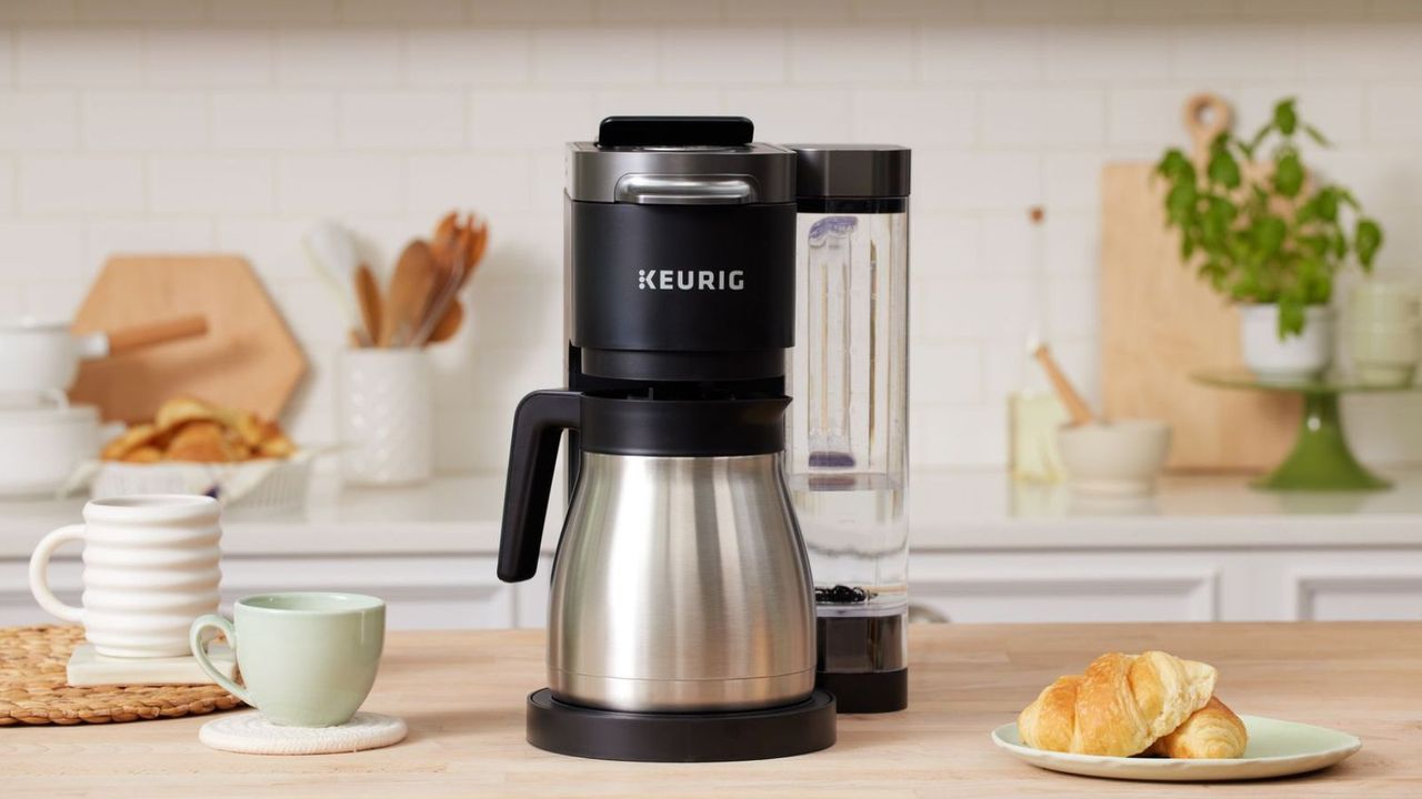 A Keurig coffee maker on a wooden countertop surrounded by mugs and a plate of pastries; in the background, kitchen utensils against a white tiled wall. 