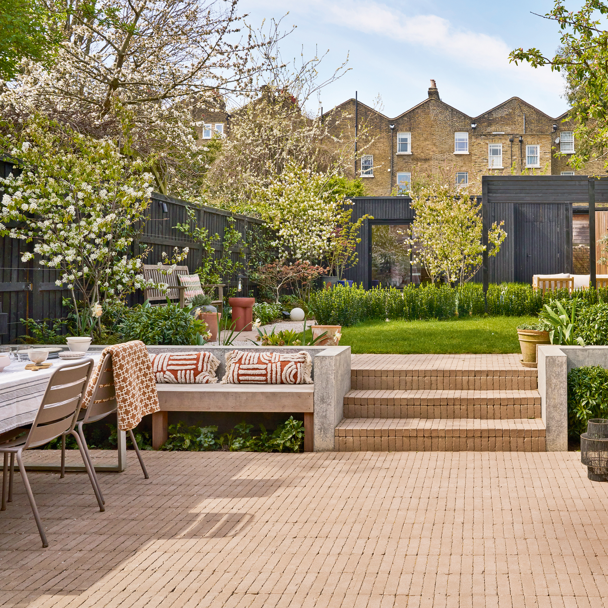 a large back garden with a black time framed studio at the end, trees and lawn in the middle and a brick patio in the foreground with a dining area