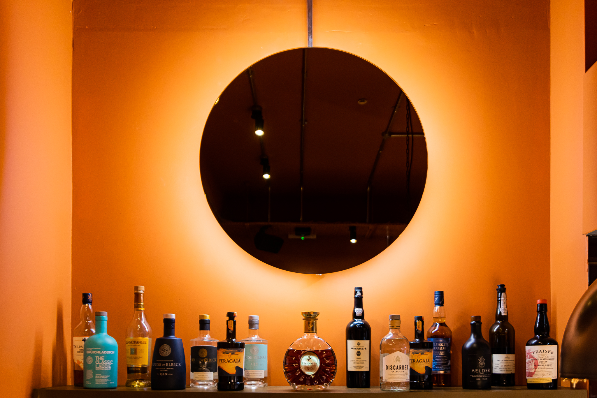 an image of the restaurant with a big circular mirror, orange walls, and a row of spirits at the bar area