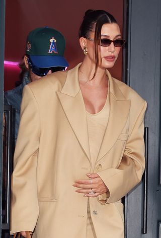 Amazon Prime dupe find of the celebrity-approved sculptural earring trend shown on a candid close photo of Hailey Bieber wearing gold bottega veneta earrings with brown sunglasses, a tan oversize blazer layered over a tan tank top dress