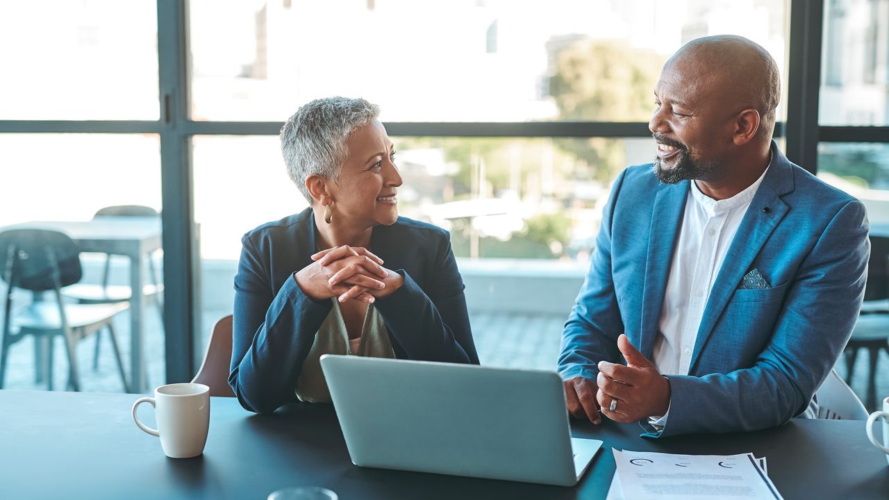 An older woman talks with a financial adviser about retirement in his office while sitting at a table with a laptop open in front of them.