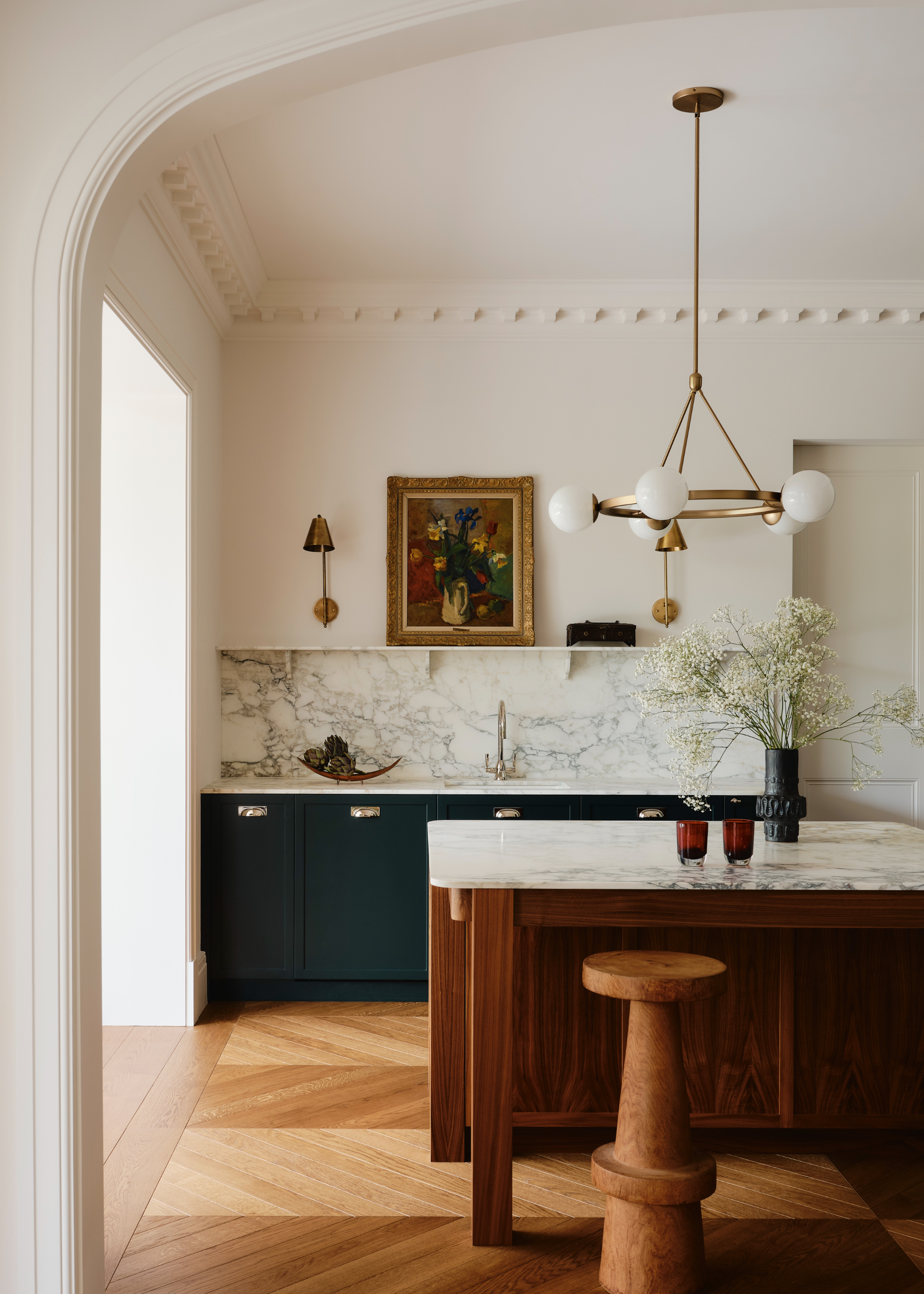 A west london kitchen with a dark wood island and a marble top, dark green cabinetry and a marble backsplash. Above the island is a gold light fixture