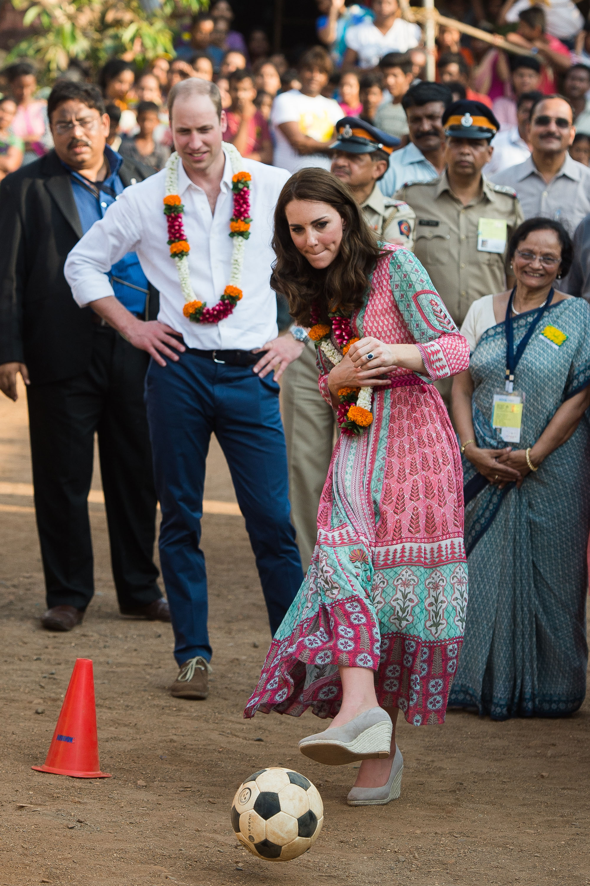 Princess Kate kicking a soccer ball wearing a dress with Prince William watching behind her