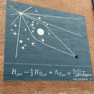 An orange brick wall with a blue square painted on it. Within the blue square is a line drawing of the gravitational lensing effect, under which is Einstein's field equation