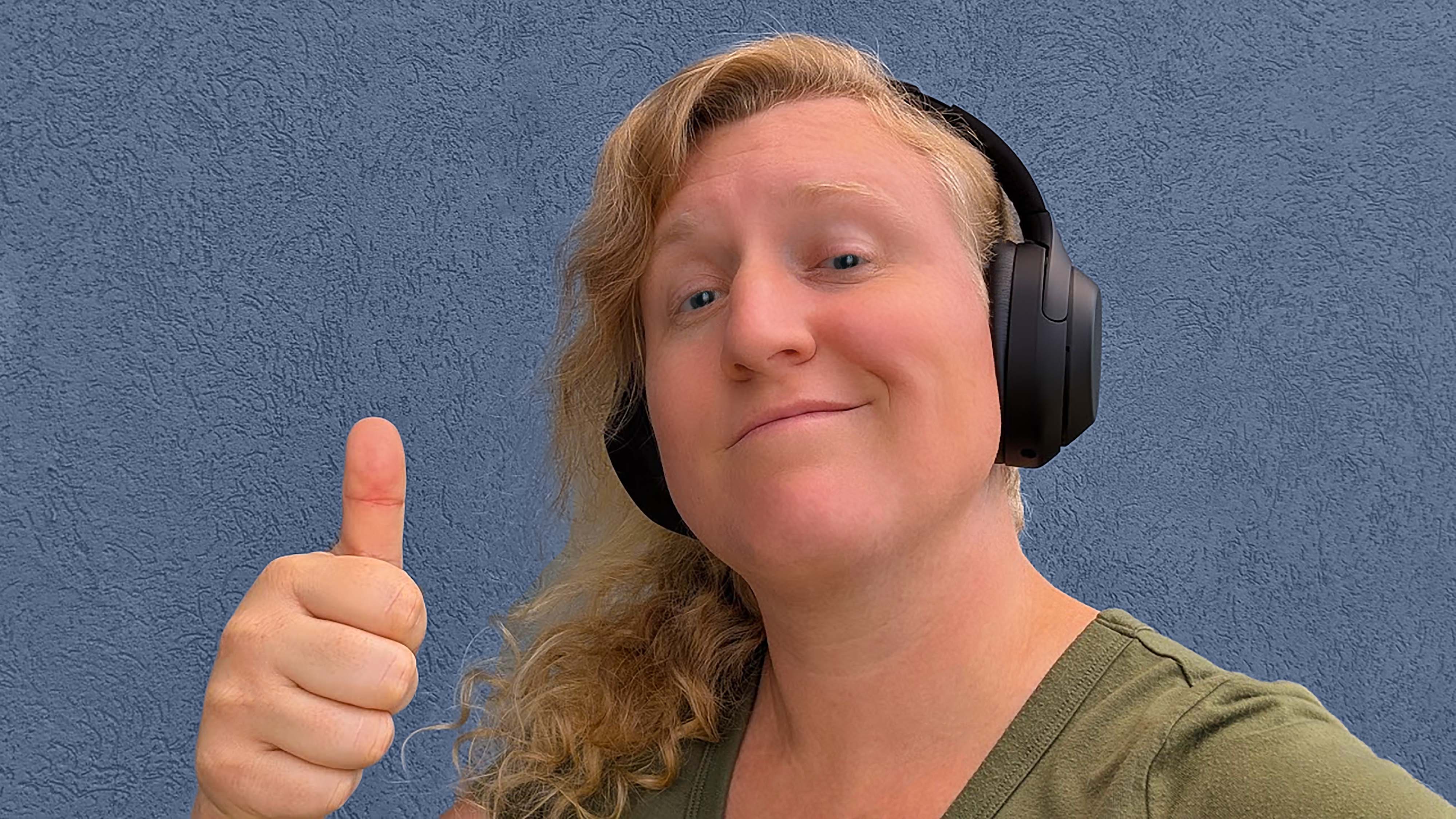 A woman wearing Sony WH-1000XM4 headphones and smiling while holding her thumb up. 