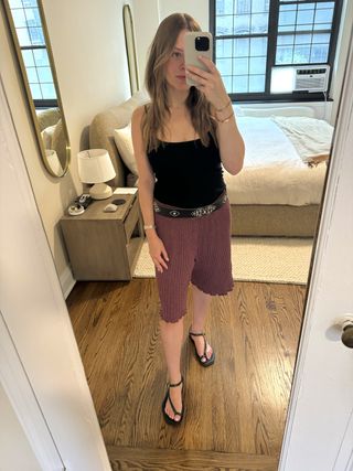 Nikki Chwatt wears Free People Jada bermuda shorts in burgundy with black sandals, a black beaded belt, and a black tank top.