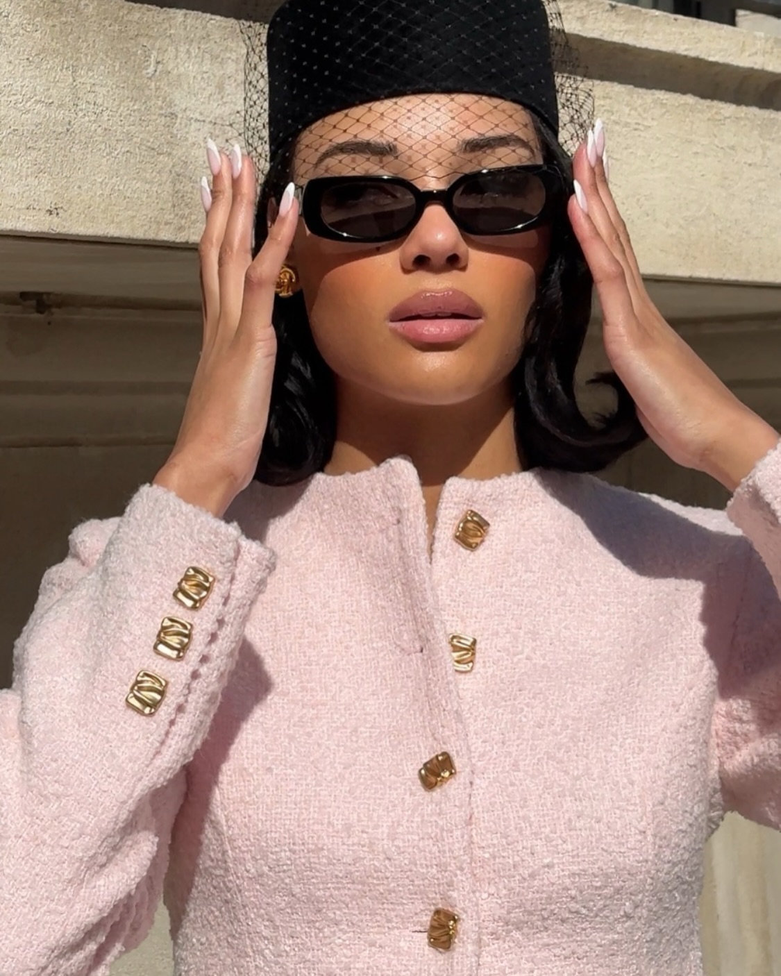 A woman wearing a pink tweed suit with a pillbox hat, an attached veil, and black sunglasses.