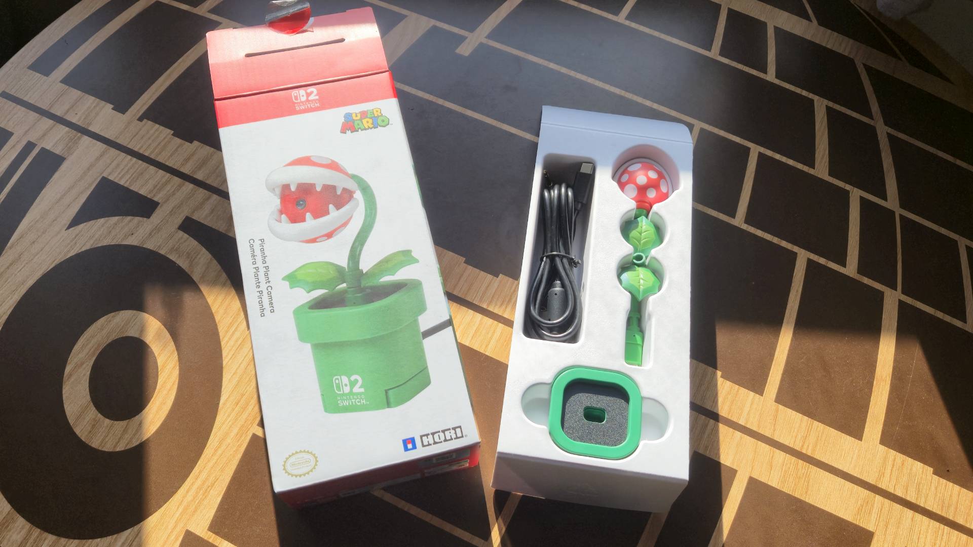 Photo by Rosalie Newcombe of the box of the Hori Piranha Plant Switch 2 camera sitting on a wooden table, with its insides to the right of it.