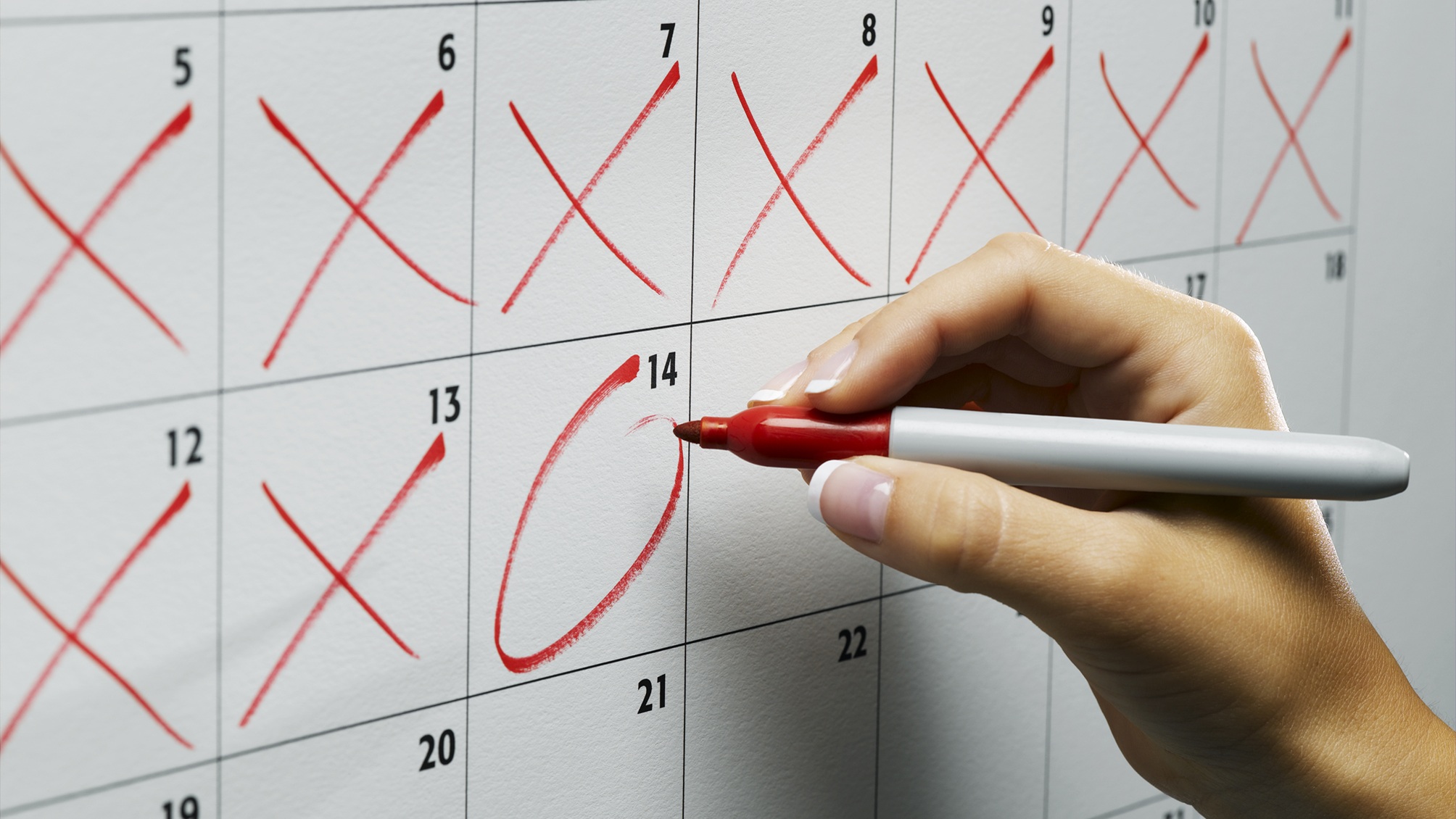 A woman circles a date on a calendar, only her hand and previous Xed-off days showing.