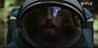 a bearded man in a spacesuit looks around confused in a forest