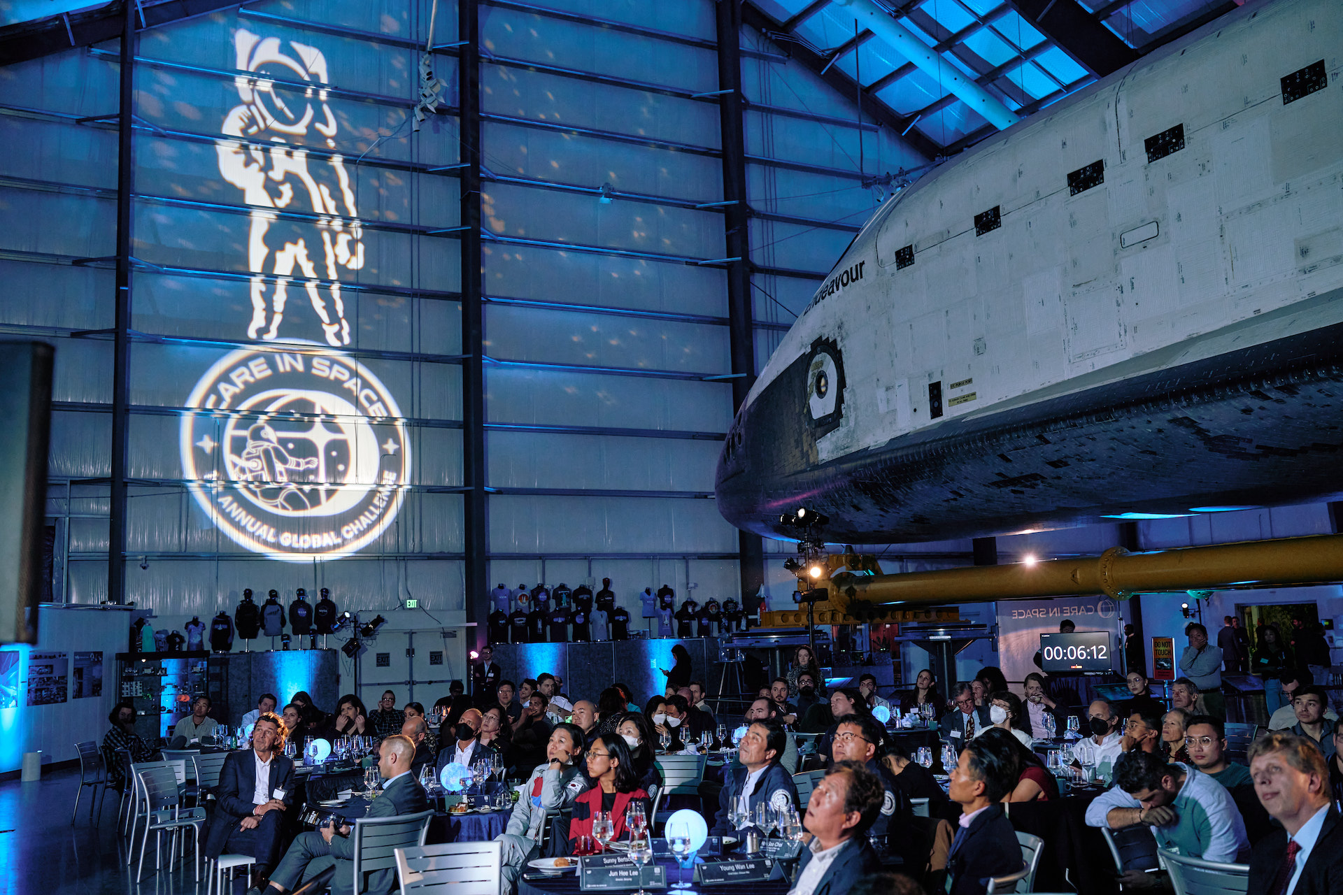 A crowd, seated around several tables, stairs up at something out of frame. Behind them, the forward body and nose of Space Shuttle Endeavour enters from the right and fills the top right quarter of the image. In the background to the left, projected against the wall, a large astronaut stands above the circular Care in Space logo patch.