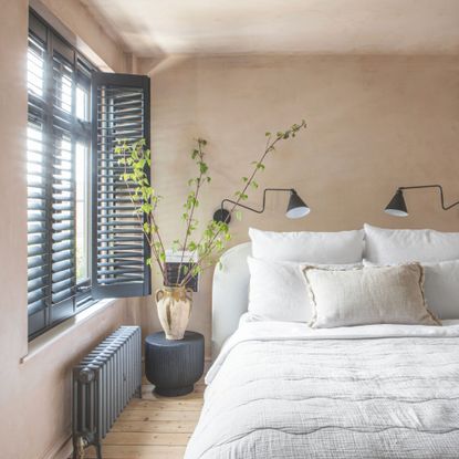 Bedroom with unfinished plaster walls, black shutters on the window on the left and a white double bed on the right