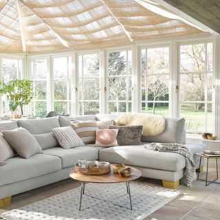 large conservatory/garden room with pale grey L-shaped sofa, blinds on ceiling, rug, small wood and metal coffee table