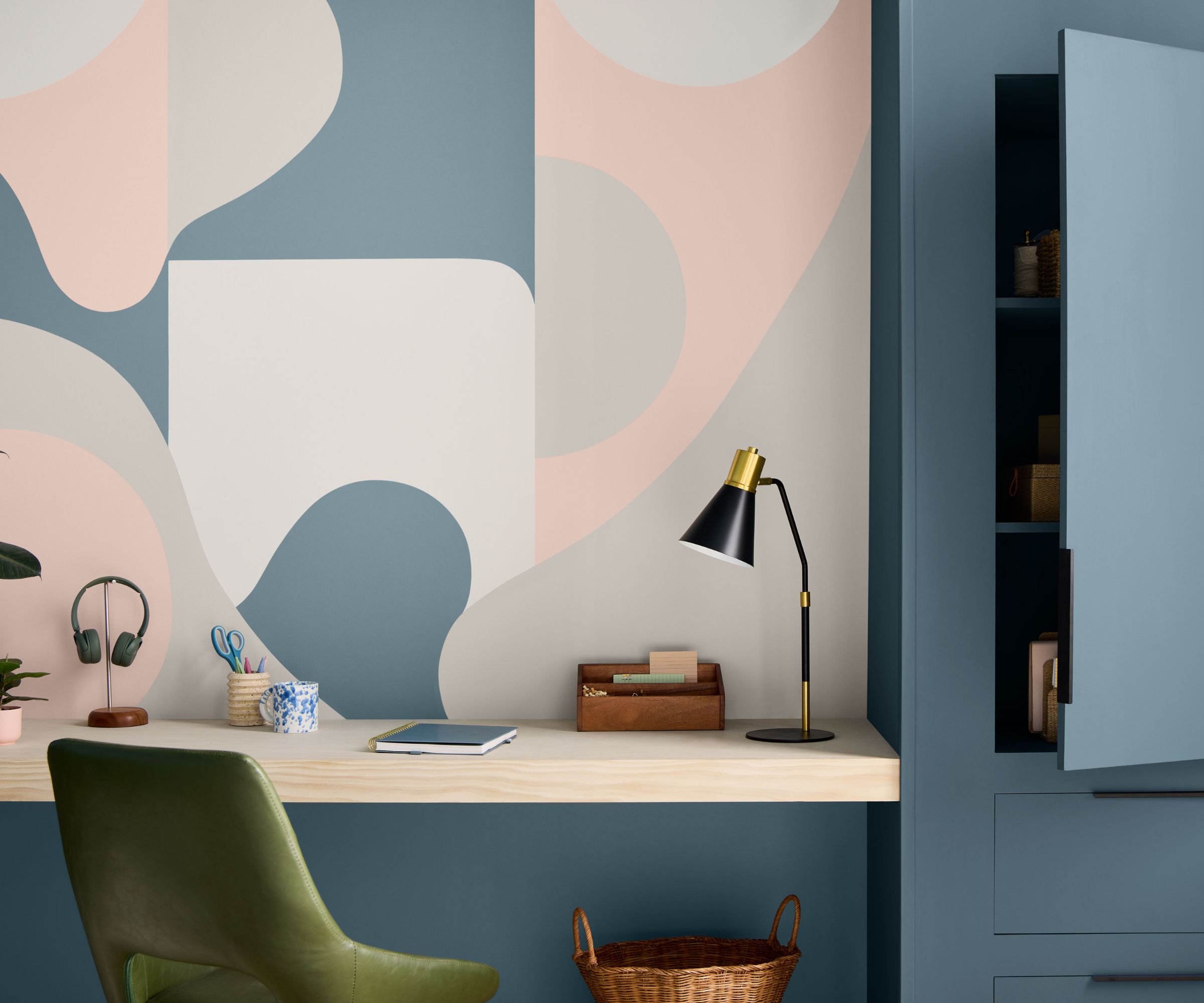 Home office painted in powder blue paint with built-in cabinetry and storage, an abstract pale pink mural, a wooden desk, a green leather desk chair, and a table lamp.