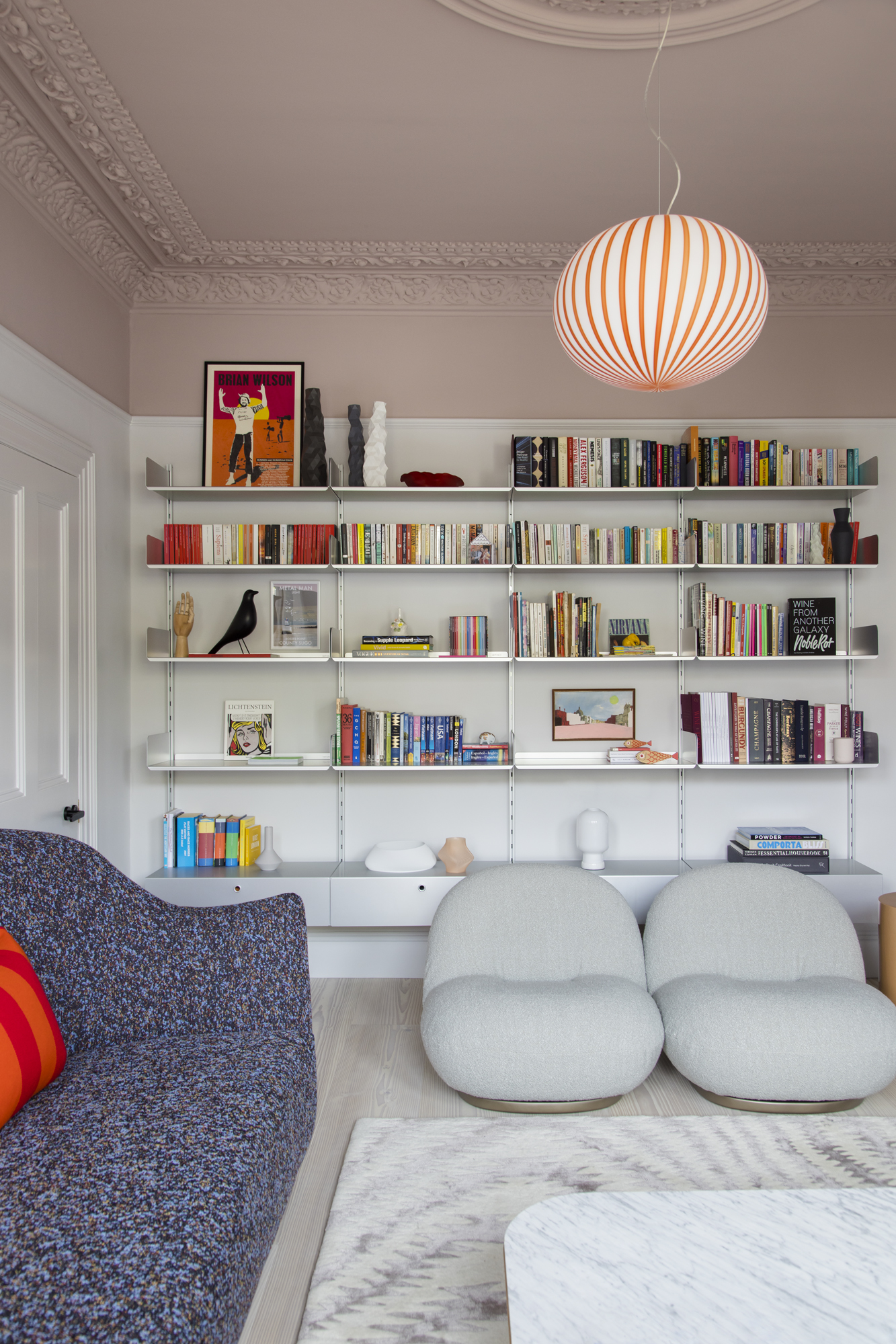 living room with floating shelves filled with books, pink ceiling with cornice, two beige armchairs, a navy sofa, a beige rug and stone coffee table and a red striped pendant light
