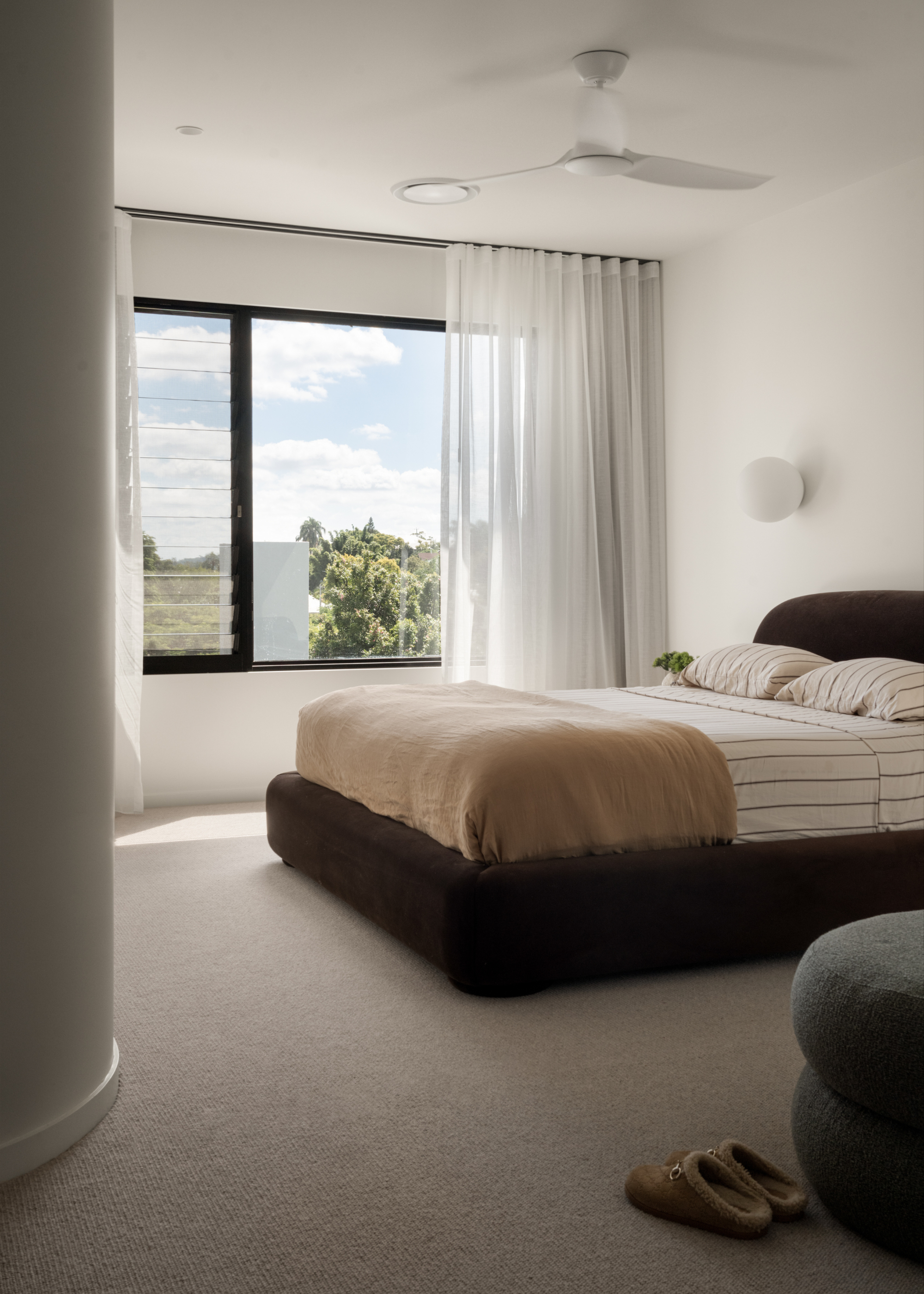 A bright white bedroom with a brown bed, striped sheets, a blue stool, and a pair of house slippers