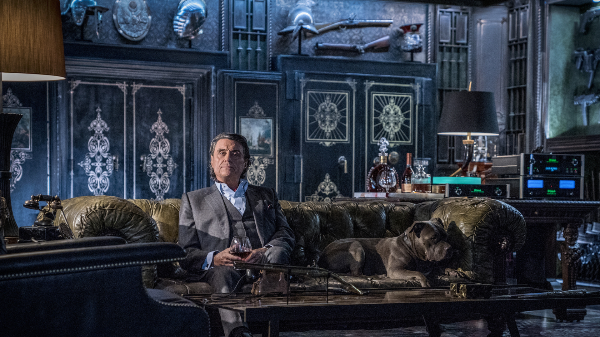 Winston smiles at someone off-camera as he sits on a fancy couch in the John Wick film series