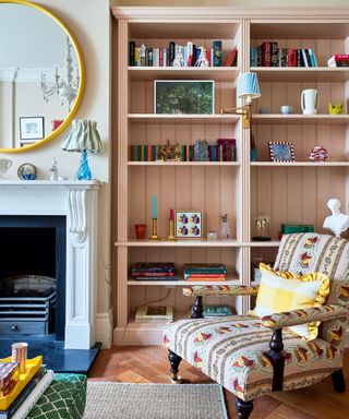 Colorful living room with bespoke pink shelving, wooden flooring, patterned upholstered armchair, fireplace with round mirror mounted above.