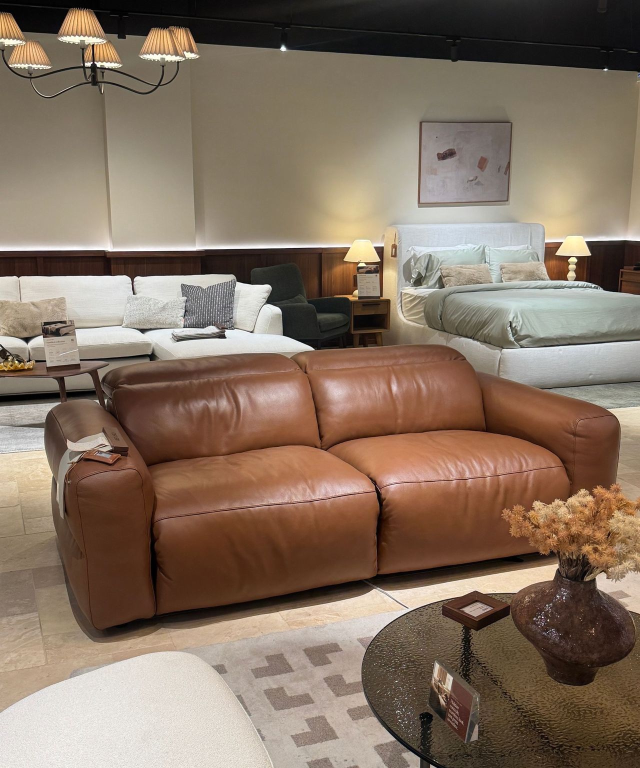 tan leather sofa on showroom floor with beds behind, glass round coffee table with vase of dried flowers