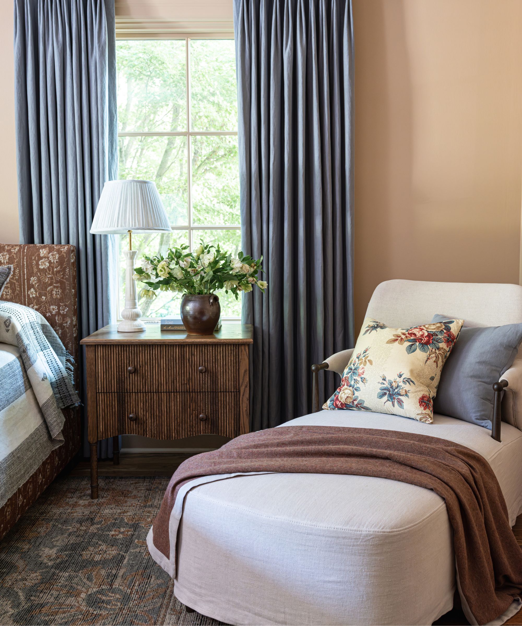 Bedroom with dusty rose walls, a Persian rug, a cream chaise lounge, a floral bed, a wooden nightstand, and blue drapes.