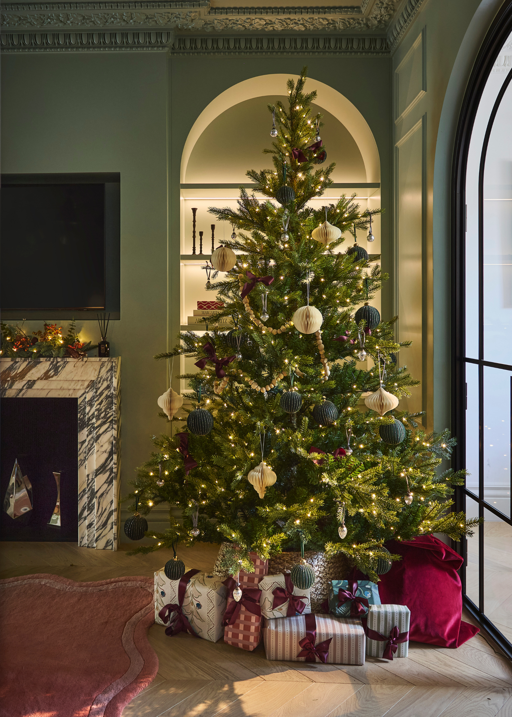 A christmas tree in front of a lit arched alcove is decorated with honeycomb decorations, lights and ribbons with presents underneath in a modern living space