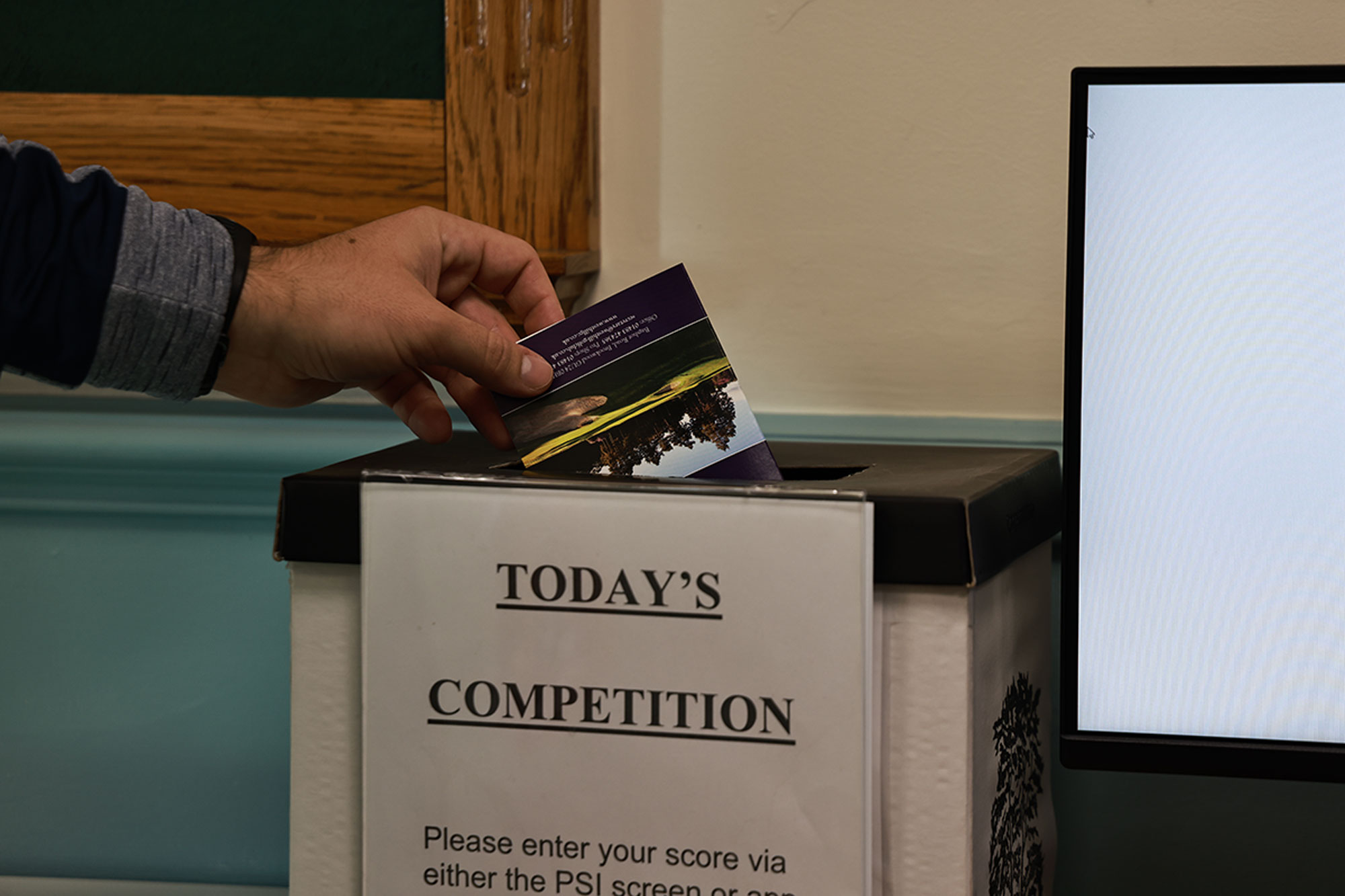 A hand placing a scorecard into a box labelled 'today's competition' in the clubhouse at a golf course