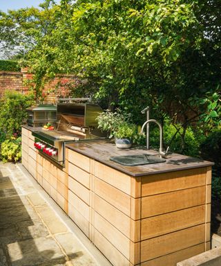 An outdoor kitchen with wooden clad side and stainless steel metalwork within a leafy garden setting