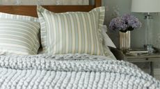 A cozy bedroom with pale blue floral wallpaper, a dark wood four-poster bed with striped cushions and grey knit throw, and a grey bedside table with silver vase and purple flowers, and lamp.