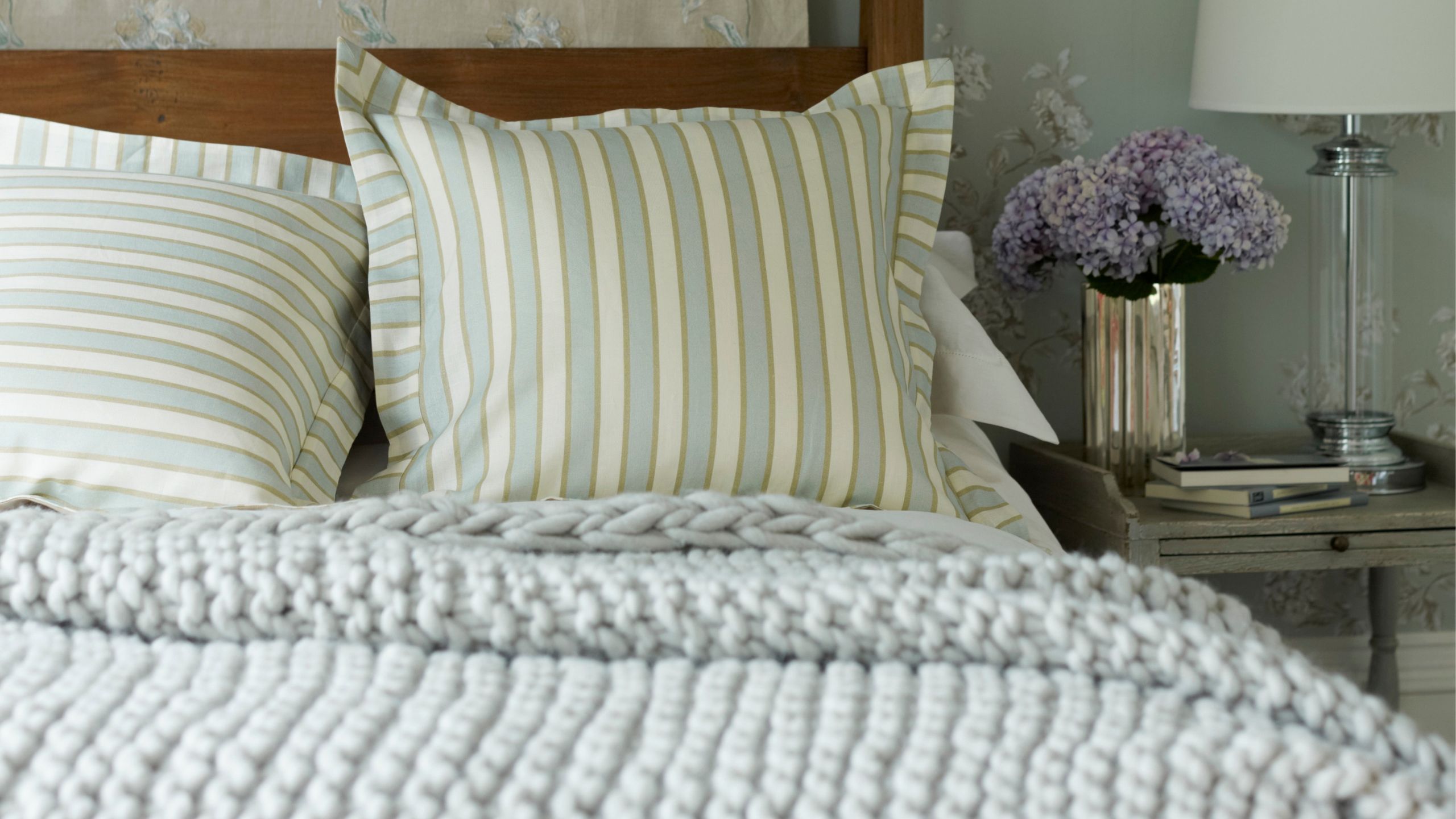 A cozy bedroom with pale blue floral wallpaper, a dark wood four-poster bed with striped cushions and grey knit throw, and a grey bedside table with silver vase and purple flowers, and lamp.