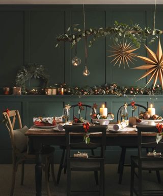 Table laid for a meal at Christmas with foliage and Christmas decoration above and Christmas decorations and lit candles in the background.