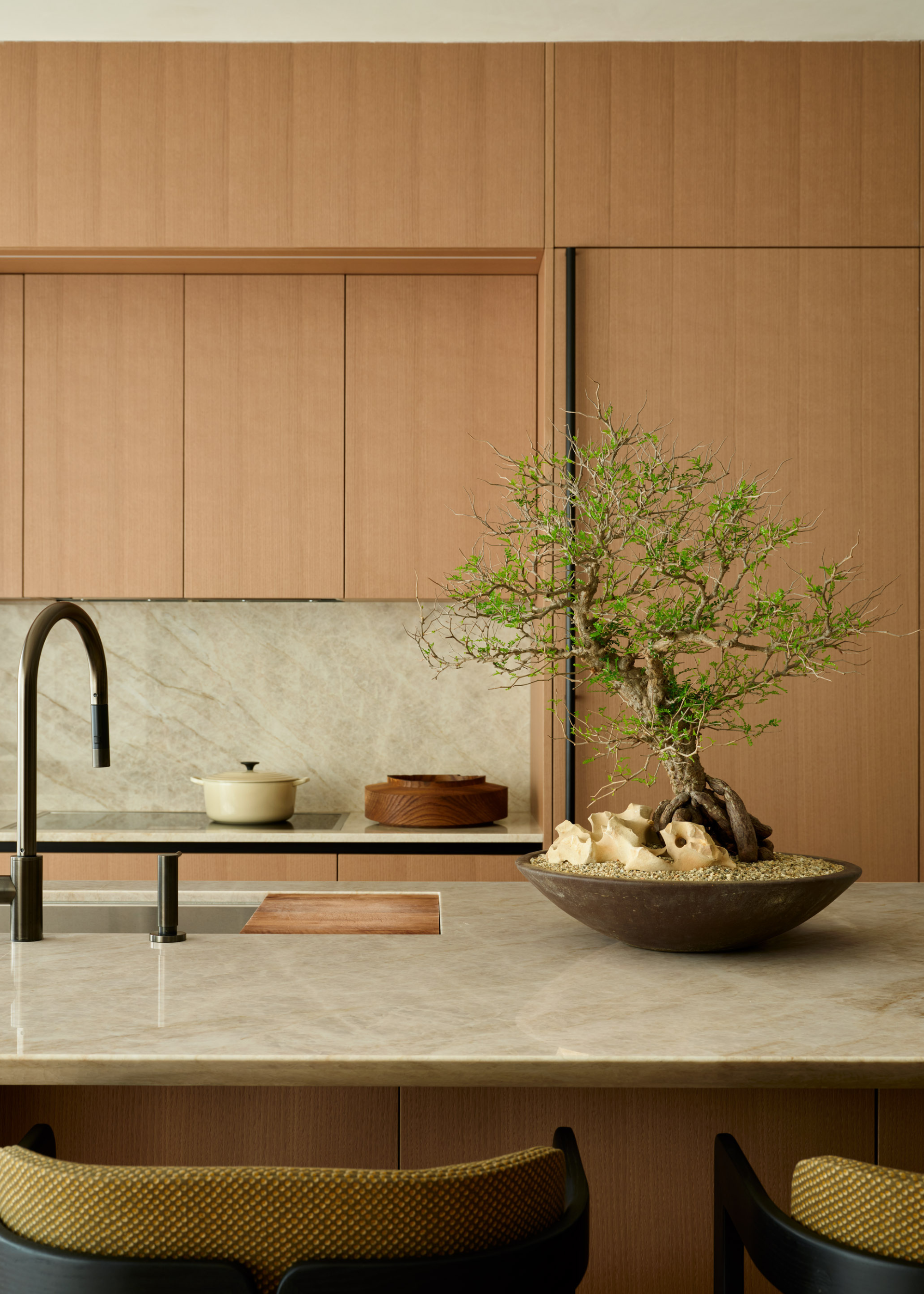 A clean kitchen with wood cabinets, a stone island, a potted bonsai, a casserole dish on the hob, and an island sink with a wood board top and black hardware by a set of green stools