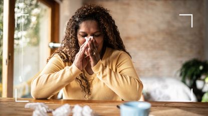 Woman using tissue to sneeze, sitting at a table at home with tissues surrounding her, after learning how to get rid of a cold fast
