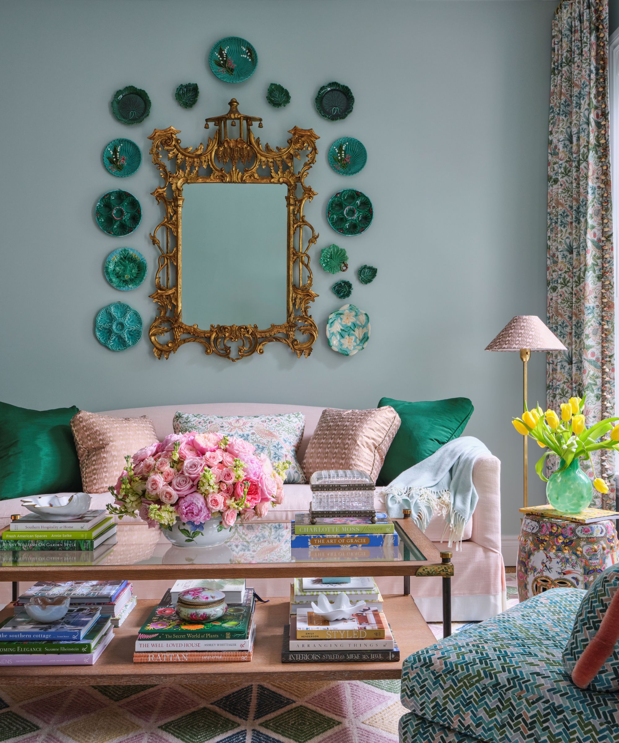 Living room with pale blue walls, a pink linen sofa decorated with teal and pink throw cushions, a large brass mirror hung on the wall above with green plates hung around it, and a glass coffee table