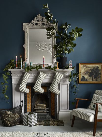 A Christmas-themed living room with navy blue wall decor, large mirror, and traditional fireplace with three white stockings