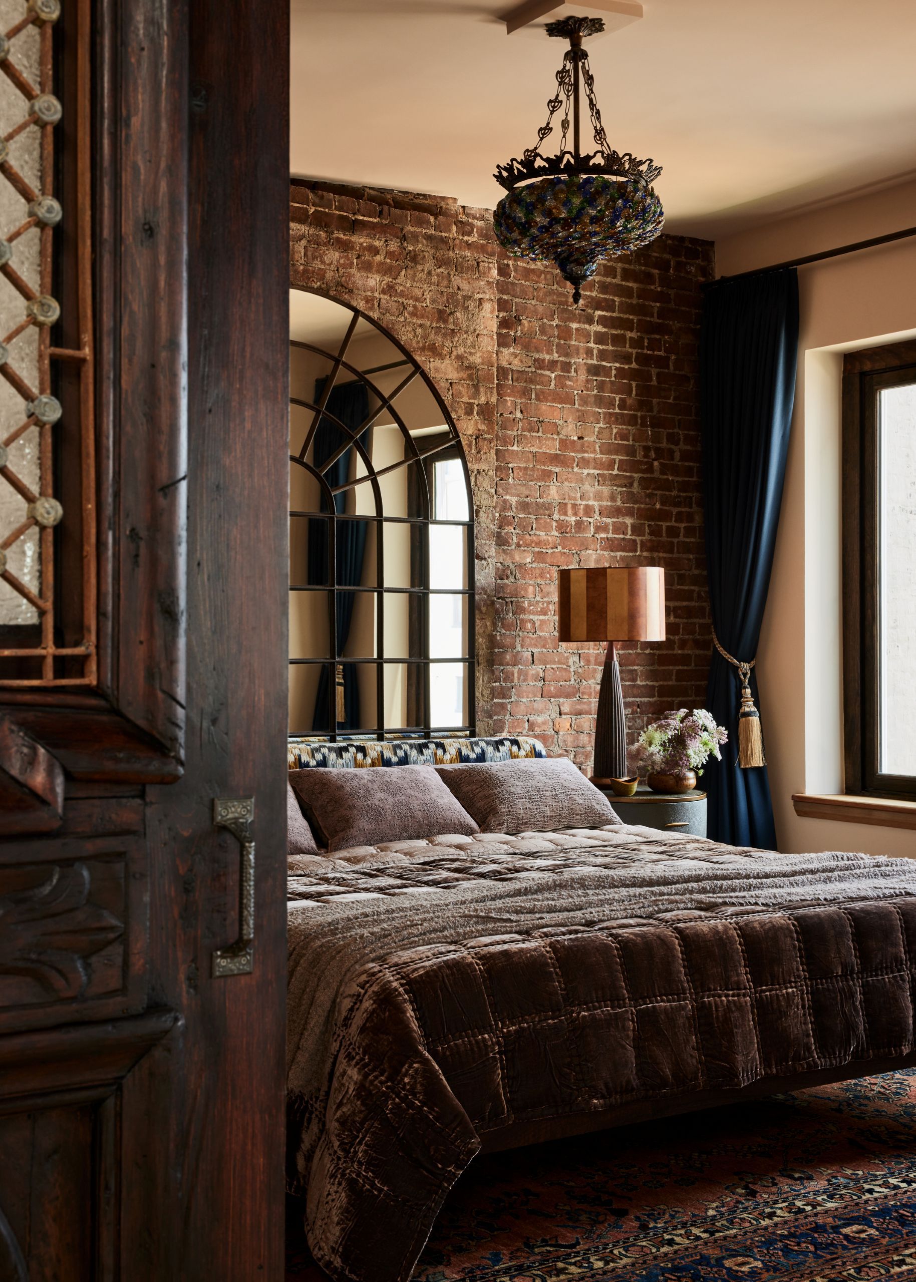 Image of a cozy bedroom with an exposed brick wall, an arched mirror behind the bed, a bed with gray velvet bedding, and a dark blue curtain with a tassel tie. 