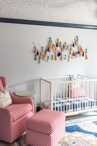 baby boy nursery with fringed wall hanging, white crib, red and white graphic print armchair and footstool, blue patterned rug, side table