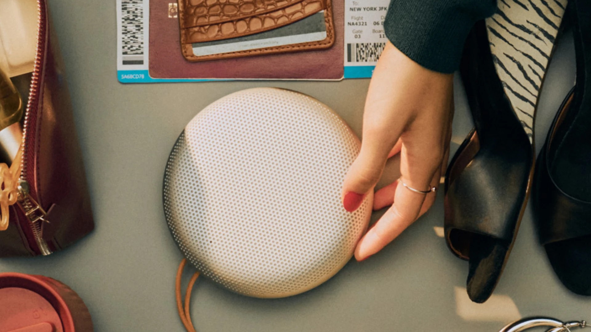 A person&#039;s hand with red nail polish rests on a round, white speaker, which is surrounded by travel items like a passport, wallet, and a flight boarding pass.