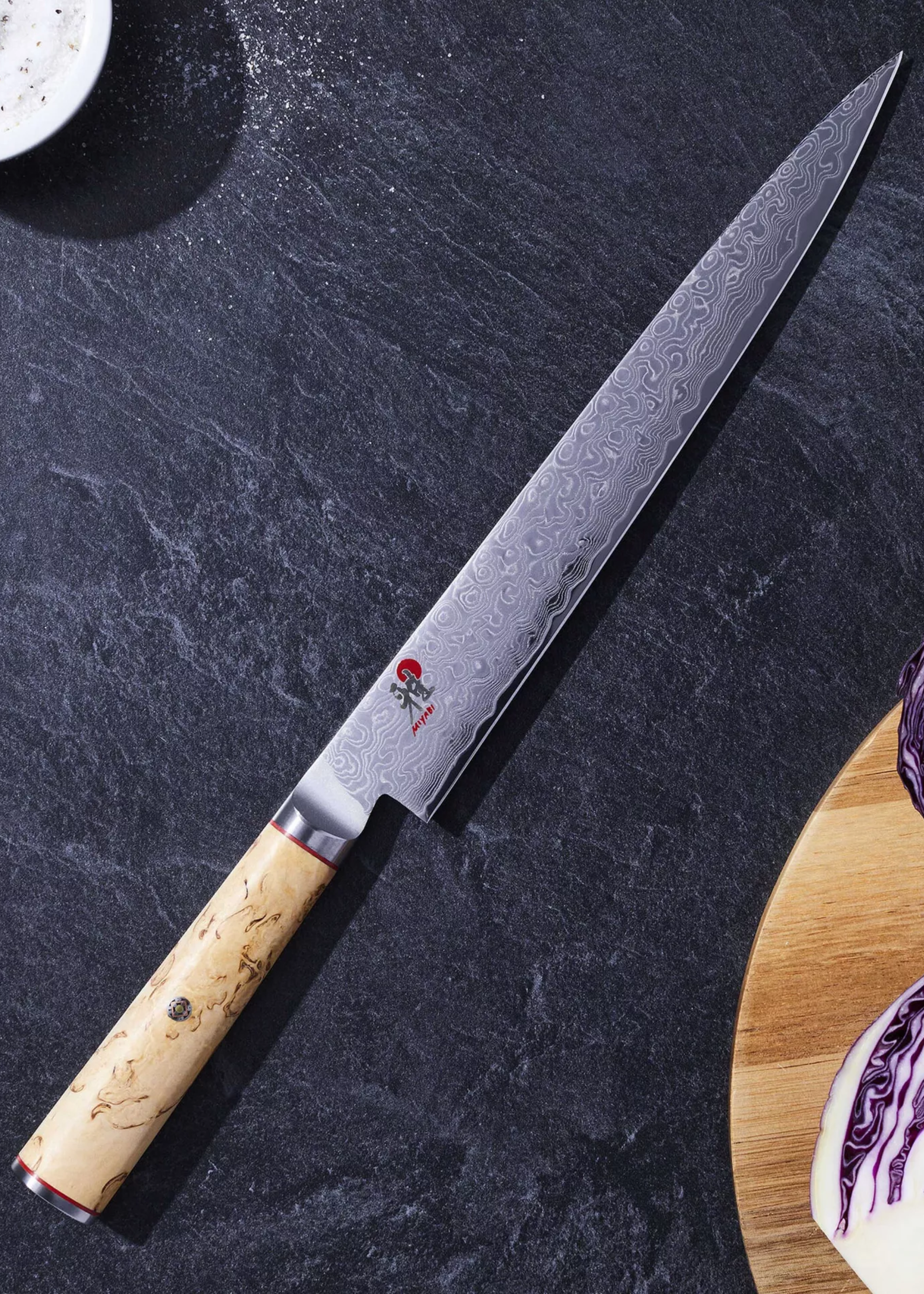 A Japanese knife on a stone countertop, near a wooden plate with a halved purple cabbage