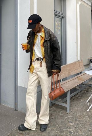 a photo showing what to wear to a hip-hop concert with a woman wearing a baseball cap with a black leather jacket layered over a yellow button-down and white t-shirt styled with white jeans, black ankle boots, and a brown bag