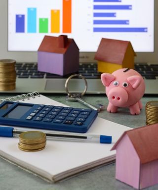 Grey surface with notepad and pen, calculator, piles of coins, piggybank, key, laptop with charts shown on screen and house models