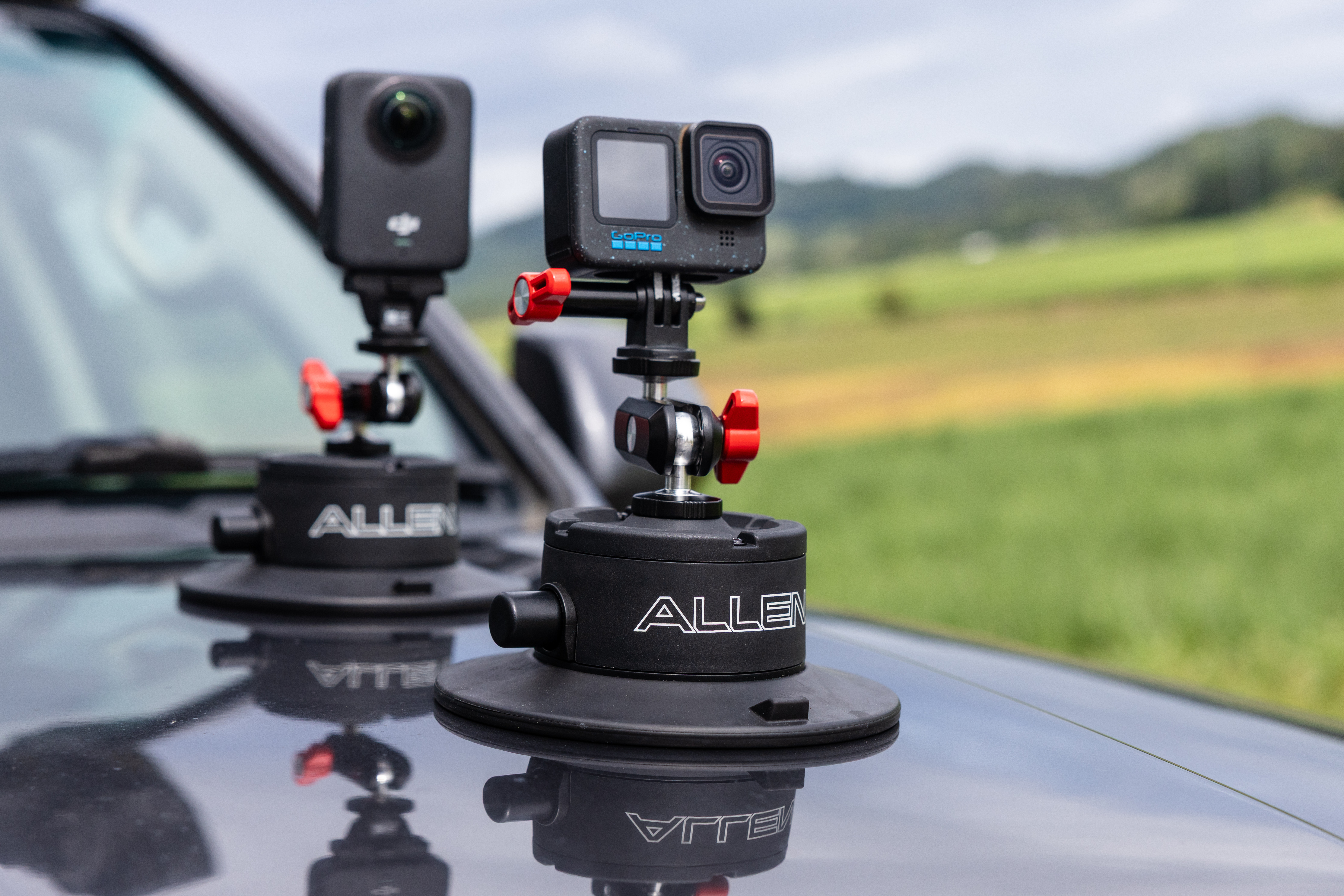 Two Allen Smart Suction Snap Camera Mounts are secured to the hood of a dark vehicle, holding a DJI action camera and a GoPro camera.